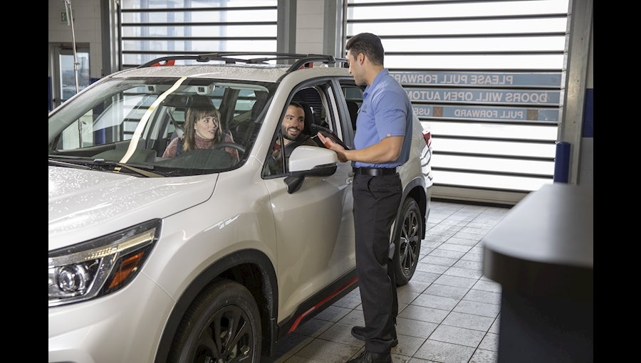 A service advisor discusses car maintenance with a couple seated in a white SUV at an auto service center.