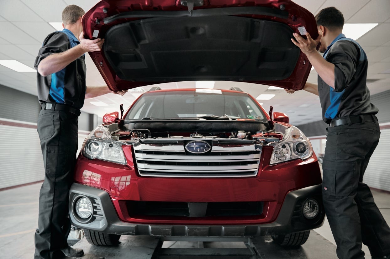 Two mechanics opening the hood of a red Subaru on a lift in a garage.