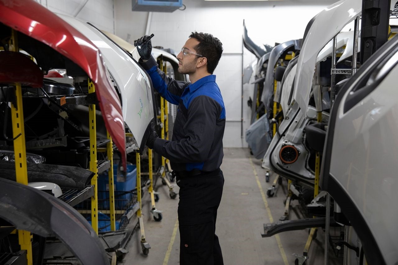 Technician wearing safety glasses and gloves inspecting a white car part with a flashlight in a warehouse.