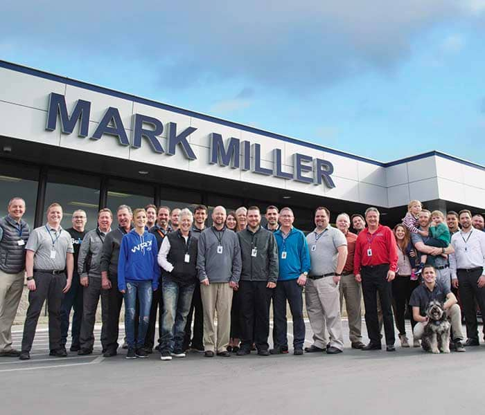 A diverse group of over 30 adults, toddlers, and a dog pose in front of a building with the 'MARK MILLER' sign.